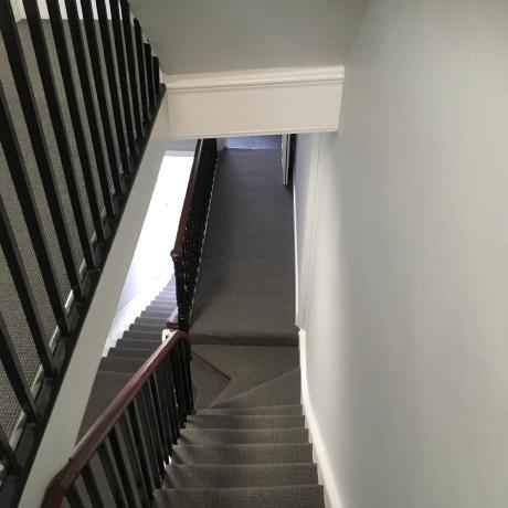 A top-down view of a staircase featuring freshly painted pale grey walls and crisp white architectural woodwork. The dark banister and charcoal grey carpet provide a modern contrast, showcasing high-end decorating work available in Islington, Covent Garden, and Canary Wharf.