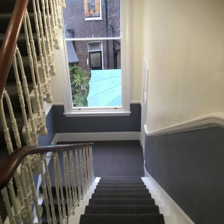 A Victorian staircase featuring professional two-tone painting with off-white upper walls and blue-grey wainscoting. The image highlights clean lines around a white sash window and ornate banisters, reflecting expert decorating for period homes in Covent Garden, Islington, and Canary Wharf.