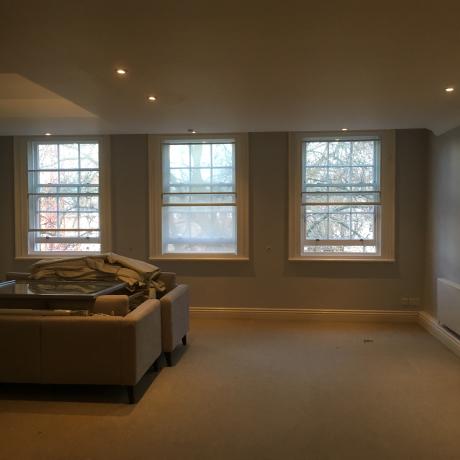 A large, modern living room with professionally painted grey walls, three white sash windows, and clean skirting boards. High-quality residential decorating service for homes in Islington, Covent Garden, and Canary Wharf.