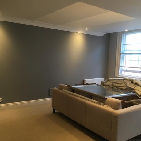 A spacious living room featuring professionally painted grey matte walls and clean white woodwork. The image shows high-quality finishes around large sash windows and decorative coving, completed by a painter and decorator serving Islington, Covent Garden, and Canary Wharf.