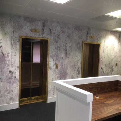 A high-end commercial lobby featuring decorative textured wall coverings around gold-framed elevator doors. The space includes a custom wood-grain reception desk and professional white trim, showcasing specialist interior decorating for offices in Canary Wharf, Islington, and Covent Garden.