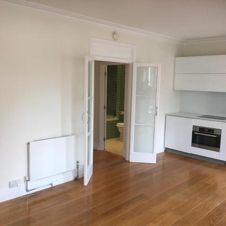 A bright, open-plan living space featuring professionally painted off-white walls and crisp white woodwork on the skirting boards and decorative coving. The room includes a modern white kitchen unit and double glass doors leading to a green-tiled bathroom, showcasing high-standard interior decorating in Islington, Covent Garden, and Canary Wharf.