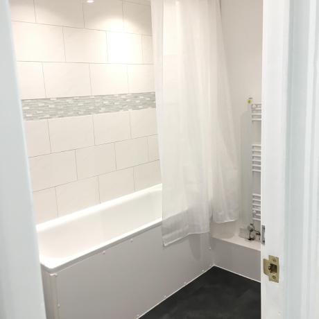 A bright, clean bathroom featuring professionally painted white walls, a white tiled bath surround with a decorative grey mosaic border, and a modern white towel radiator. The high-quality finish on the woodwork and ceiling showcases expert decorating services in Islington, Canary Wharf, and Covent Garden.