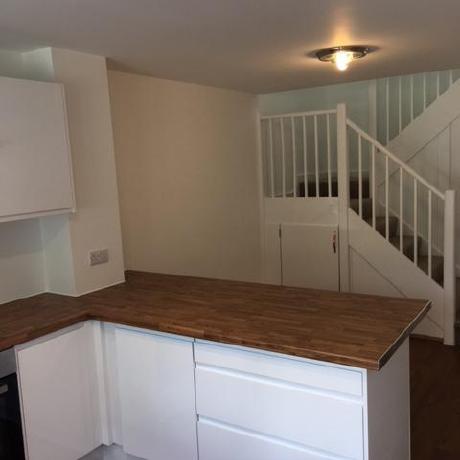 A modern white kitchen featuring a natural wood-effect countertop and professionally painted white walls. The background shows a clean, white-painted staircase and banister, showcasing a high-quality residential finish by a professional decorator in Islington, Covent Garden, and Canary Wharf.