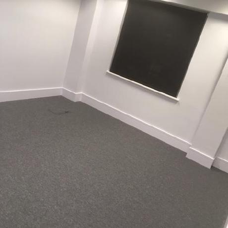 A professionally decorated office space featuring crisp white painted walls and matching white skirting boards. The image shows high-quality finishes around a large window with a dark roller blind and new grey carpeting, highlighting expert commercial painting services in Canary Wharf, Islington, and Covent Garden.