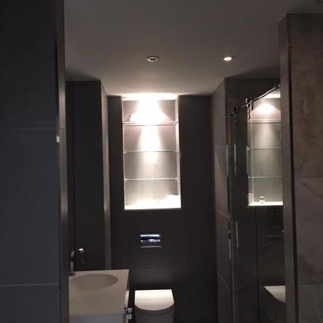 A sleek, modern bathroom featuring dark grey tiled walls and integrated glass shelving with soft recessed lighting. The space includes a contemporary white vanity and wall-hung toilet, reflecting high-end interior finishing in Canary Wharf, Islington, and Covent Garden.