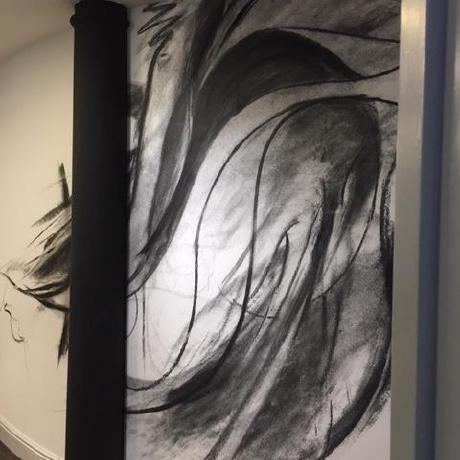 A large-scale, expressive black-and-white charcoal-style wall mural in a modern interior. The artwork is framed by a minimalist black pillar and clean white skirting boards, showcasing bespoke decorative wall-finishing services in Islington, Covent Garden, and Canary Wharf.