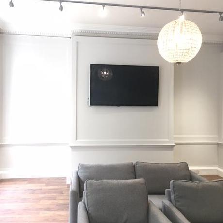 A modern living room featuring professionally painted light grey walls with decorative wall panelling and a crisp white dado rail. The space includes grey seating and contemporary track lighting, showcasing high-quality interior decorating and wood trim finishing
