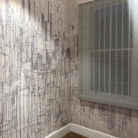 A room featuring a detailed architectural cityscape mural on the walls, paired with a white skirting board and traditional oak parquet flooring. The image showcases expert wallpaper hanging and interior decorating services for modern homes