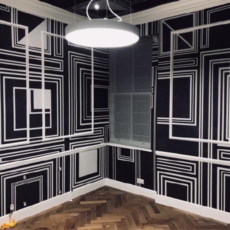 A striking room featuring a bold black and white geometric pattern on the walls and ceiling, accented by white decorative wall panelling. The space includes high-end oak parquet flooring and a modern circular pendant light, showcasing specialist luxury decorating and wallpapering services