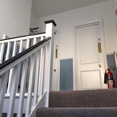 A view looking up a carpeted staircase in a period property, featuring a white-painted banister with a dark contrasting handrail. The hallway showcases two-tone walls with a soft blue lower section and crisp white upper walls and doors, reflecting professional interior decorating and period restoration in Islington, Covent Garden, and Canary Wharf.