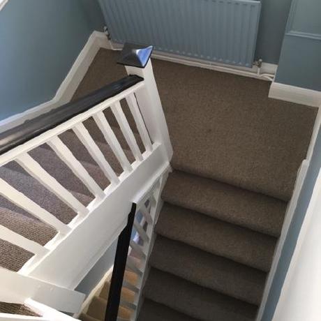 A top-down view of a professionally decorated period staircase featuring a white-painted banister with a dark handrail and grey carpeting. The hallway walls are finished in a two-tone style with a soft blue lower section and a matching blue radiator, showcasing high-end interior decorating