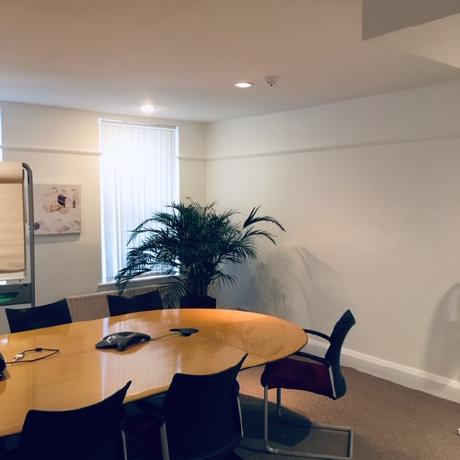 A bright, professionally decorated meeting room featuring crisp white painted walls and matching white skirting boards. The space includes a large wood-finish conference table, black office chairs, and integrated ceiling lighting, showcasing expert commercial interior finishing in Canary Wharf, Islington, and Covent Garden.