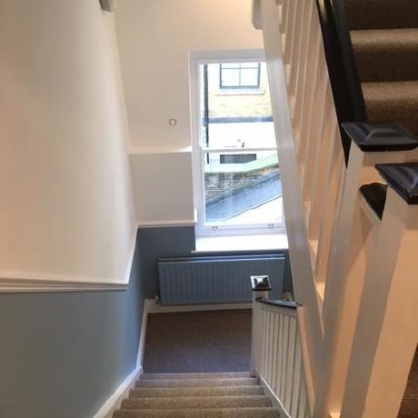 A top-down view of a professionally decorated period staircase featuring a clean white banister with a dark handrail and grey carpeting. The hallway showcases a two-tone wall finish with a soft blue lower section, crisp white upper walls, and a matching blue radiator, reflecting high-quality residential decorating in Islington, Covent Garden, and Canary Wharf.