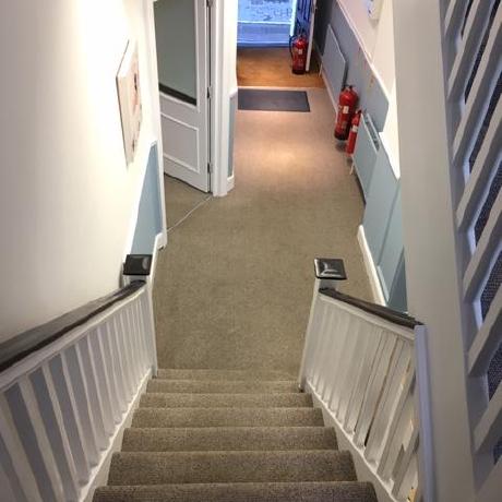 A view looking down a carpeted staircase in a period property, showing a light grey carpeted hallway with a two-tone wall finish in soft blue and white. The space features a white banister with a dark handrail, a matching blue radiator, and professionally painted white doors, showcasing expert residential decorating