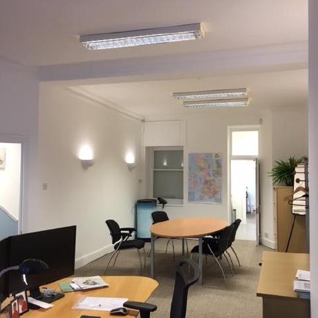 A bright, open-plan office space featuring professionally painted white walls and crisp white ceiling finishes. The room includes multiple workstations, a central meeting table, and integrated lighting, showcasing expert commercial painting and decorating services in Canary Wharf, Islington, and Covent Garden.