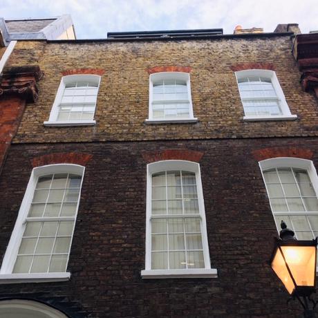 A low-angle view of a brick period building featuring six professionally painted white sash windows with arched brick headers. The image showcases high-quality exterior maintenance and precision woodwork painting in Covent Garden, Islington, and Canary Wharf.