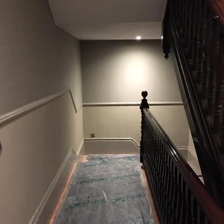 A period hallway featuring professionally painted taupe walls with matching decorative dado rails and skirting boards. An ornate dark wood banister is prominent, and the floor is covered with Sticky Shield protective film to ensure a clean work environment during decorating services in Islington, Covent Garden, and Canary Wharf.