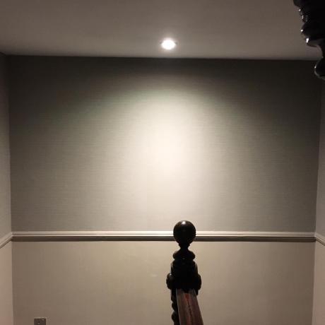 A close-up view of a professionally decorated period wall featuring a subtle textured wallpaper above a clean taupe-painted lower section. The space is divided by a perfectly finished dado rail and accented by a dark wood banister finial, showcasing expert interior decorating for period homes