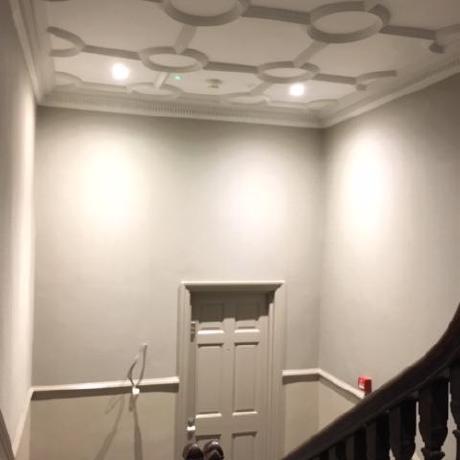 A high-angle view of a professionally decorated period hallway featuring an ornate white plasterwork ceiling with a geometric circular design. The walls are finished in a two-tone neutral palette with a decorative dado rail, and the space includes a matching painted door and a dark wood banister, showcasing expert heritage interior work in Islington, Covent Garden, and Canary Wharf.