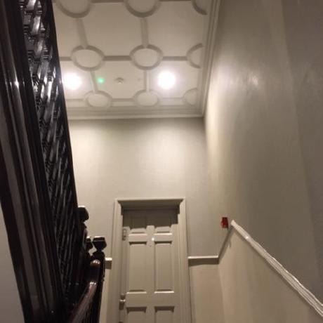 A low-angle view looking up a period staircase toward an ornate white plasterwork ceiling featuring a geometric circular pattern. The hallway showcases professionally painted walls in a two-tone neutral palette with a decorative dado rail, matching door, and a dark wood banister, highlighting specialized interior redecoration in Islington, Covent Garden, and Canary Wharf.