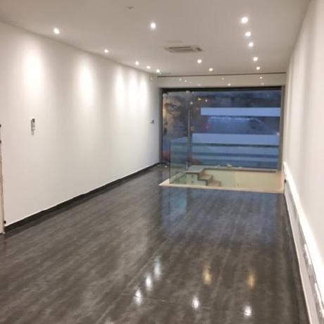 A long, modern commercial interior featuring professionally painted white walls and a high-gloss grey tiled floor. The space includes recessed spotlighting and a glass-enclosed stairwell, showcasing high-end commercial finishing for retail or office spaces in Canary Wharf, Islington, and Covent Garden.
