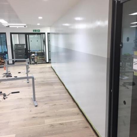 A modern office interior under renovation featuring a professionally painted two-tone feature wall in white and soft grey. The space includes light wood flooring, glass partitions, and recessed LED lighting, showcasing high-standard commercial decorating in Canary Wharf, Islington, and Covent Garden.