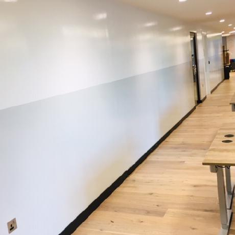 long perspective view of a modern office hallway featuring a professionally painted high-gloss two-tone feature wall in white and pale grey. The space is finished with light wood flooring, black skirting boards, and recessed ceiling spotlights, showcasing high-end commercial decorating services in Canary Wharf, Islington, and Covent Garden.