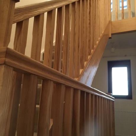 A low-angle view of a contemporary staircase featuring solid oak spindles, handrails, and newel posts with a clear protective finish. The space is accented by neutral-painted walls and natural light from a nearby window, showcasing expert wood treatment and interior decorating in Islington, Covent Garden, and Canary Wharf.