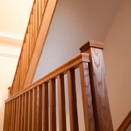 A low-angle shot of a contemporary staircase featuring vertical oak spindles and a matching square newel post with a visible natural wood grain. The woodwork is professionally finished to a smooth sheen, complemented by neutral-toned walls, highlighting high-quality carpentry and decorating services in Islington, Covent Garden, and Canary Wharf.