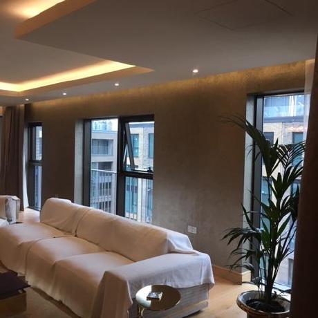 A modern luxury living room featuring a textured gold or bronze metallic feature wall and a recessed tray ceiling with integrated LED strip lighting and spotlights. The space is finished with light oak flooring and large floor-to-ceiling windows, showcasing premium interior decorating